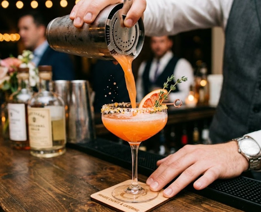 Professional bartender pouring a salted-rim craft cocktail from a shaker into a coupe glass at a private event